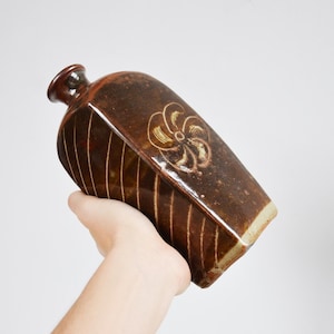 May include: A brown ceramic bottle with a flower design in a lighter brown color. The bottle has a slightly rounded shape and a narrow neck.