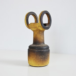 May include: A yellow and black ceramic vase with two circular openings at the top. The vase has a textured surface and a unique design.