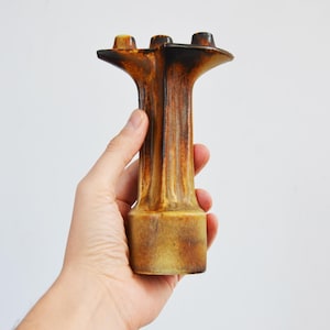 May include: A brown and tan ceramic candle holder with three openings. The candle holder has a textured, vertical design and is held in a person's hand against a white background. The top of the candle holder is dark brown.