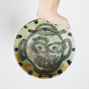 May include: A ceramic bowl with a hand-painted design of a face. The face is painted in shades of brown, blue, and green. The bowl has a white background.