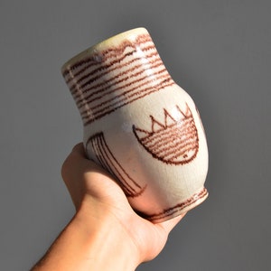 May include: A white ceramic vase with brown painted lines and a stylized face design.