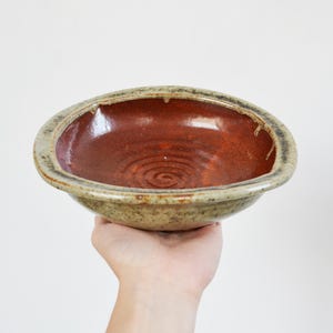 May include: A round ceramic bowl featuring a glossy, deep reddish-brown interior with a visible spiral pattern at the base. The exterior rim and sides are finished in a speckled, muted beige and grey glaze with a textured, earthy appearance.