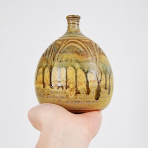 Può includere: Un vaso in ceramica fatto a mano con un corpo arrotondato e un collo stretto. Il vaso presenta una miscela di tonalità terrose, tra cui marrone, giallo e verde, con un effetto smaltato a goccia. Il vaso è tenuto in una mano.