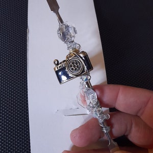 Pode incluir: Uma ferramenta prateada com um amuleto de câmera preto e contas de cristal transparentes. O amuleto da câmera tem o texto "FM2" e "Glider".