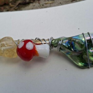 May include: A handcrafted hair accessory. It features a yellow crystal, a red and white mushroom bead, and a green iridescent glass piece, all wrapped with silver wire on a gold-coloured metal stick.