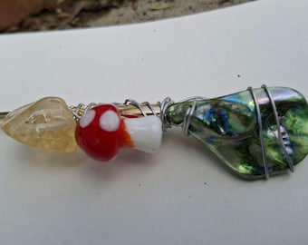 Può includere: Accessorio per capelli fatto a mano con un cristallo giallo, una perlina a forma di fungo rosso e bianco e un pezzo di vetro verde iridescente, il tutto avvolto con filo d'argento su un bastoncino di metallo dorato.