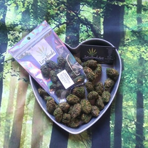 May include: A heart-shaped gray dish filled with small, green, round cannabis buds. A clear plastic bag with the text "Love Buds" is on top of the dish. A business card with the text "Love Buds.Art" is visible in the background.