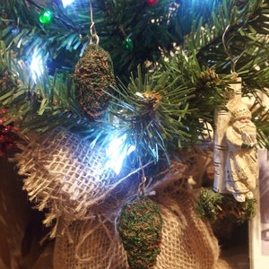 Könnte beinhalten: Ein kleiner künstlicher Weihnachtsbaum, geschmückt mit grünen Tannenzapfen-Ornamenten, weißen Lichtern und einem Jutesack an der Basis.