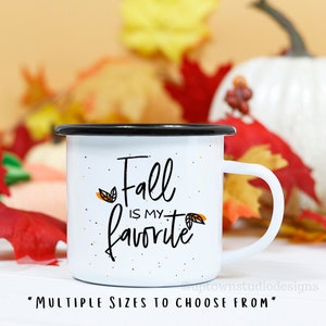 May include: White enamel mug with a black rim and handle. The mug has a white background with black and orange text that reads "Fall is my favorite". The mug is surrounded by fall leaves.