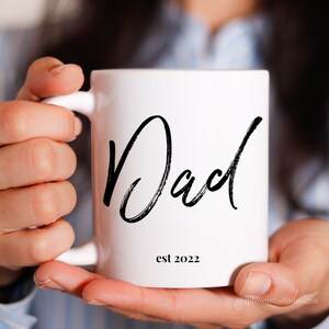 May include: White ceramic mug with a black handwritten inscription that reads "Dad est 2022".