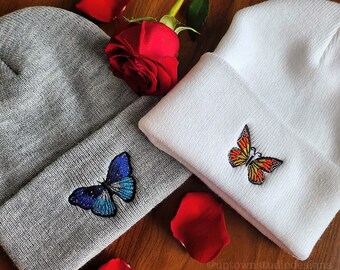 May include: Two knit beanies with embroidered butterfly designs. One beanie is gray with a blue butterfly, the other is white with an orange butterfly.