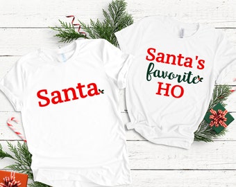 Può includere: Due magliette bianche con testo natalizio rosso e verde. Una maglietta dice "Santa" e l'altra dice "Santa's favorite HO".