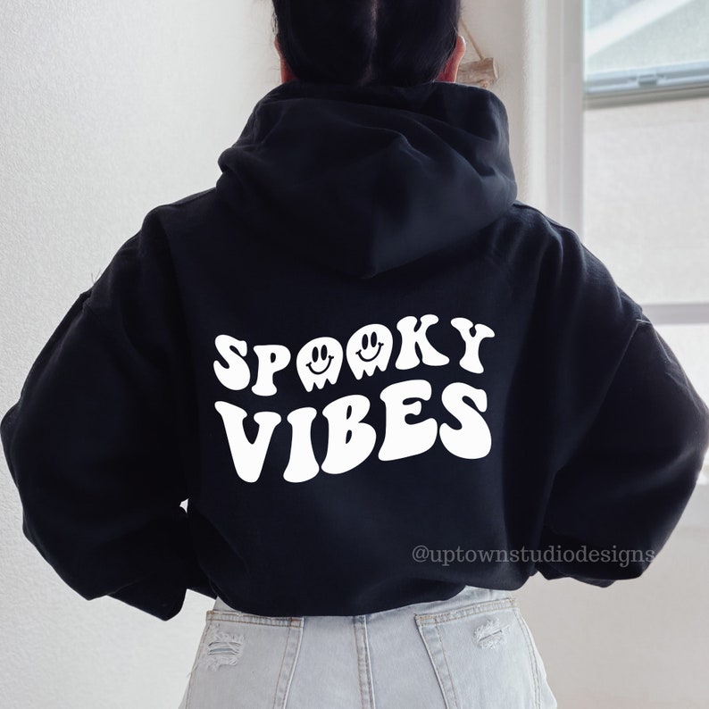 May include: Black hooded sweatshirt with white text that reads "Spooky Vibes" in a retro font. Two white ghost faces with smiley faces are above the text. The text "@uptownstudiodesigns" is at the bottom of the sweatshirt.