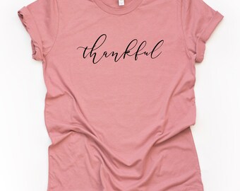Puede incluir: Una camiseta de color rosa polvoriento con la palabra "agradecido" escrita en cursiva negra.