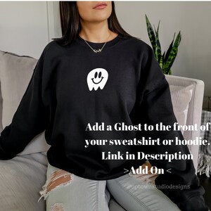 May include: Black sweatshirt with a white smiling ghost graphic on the front. The text "Add a Ghost to the front of your sweatshirt or hoodie. Link in Description >Add On< @uptownstudiodesigns" is printed below the graphic.
