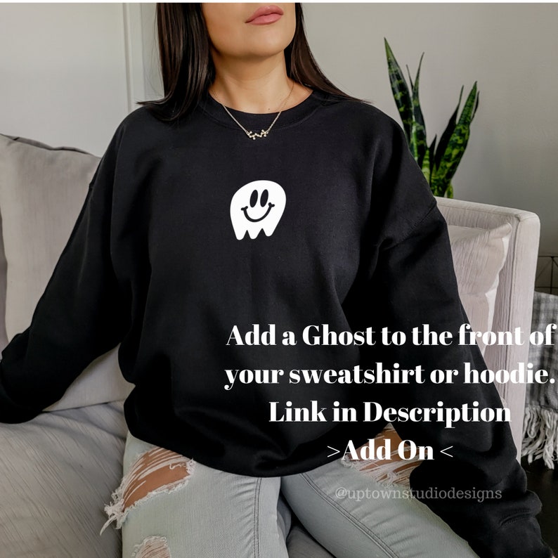 May include: Black sweatshirt with a white smiling ghost graphic on the front. The text "Add a Ghost to the front of your sweatshirt or hoodie. Link in Description >Add On< @uptownstudiodesigns" is printed below the graphic.