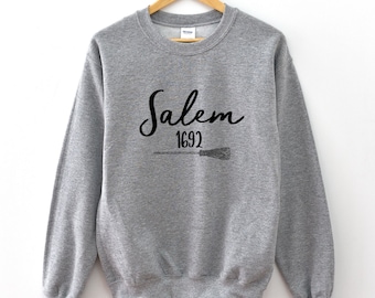 Op de afbeelding: Grijs sweatshirt met zwarte tekst die "Salem 1692" zegt en een zwarte bezem afbeelding.