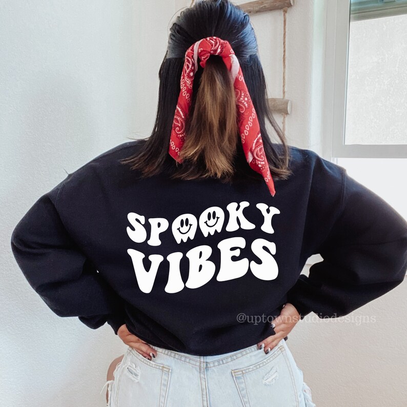 May include: Black sweatshirt with white text that reads "Spooky Vibes" in a retro font. The text is accompanied by three white ghost silhouettes with smiling faces.