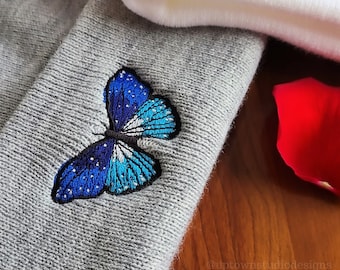 May include: A gray beanie with a blue and white butterfly embroidered on the side. The beanie is lying on a wooden surface with rose petals.