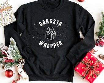 Pode incluir: Camisola de moletom preta com texto branco que diz "Gangsta Wrapper" e um contorno branco de uma caixa de presente com um laço. A camisola está cercada por decorações de Natal.