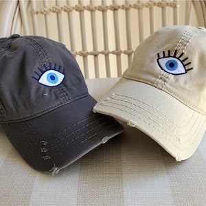 Puede incluir: Dos gorras de béisbol de algodón con un diseño de ojo azul malvado. Una gorra es gris oscuro y la otra es beige claro.