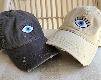 May include: Two cotton baseball caps with a blue evil eye design. One cap is dark gray and the other is light beige.