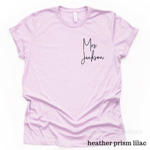May include: A light purple t-shirt with the text "Mrs. Jackson" in black script.