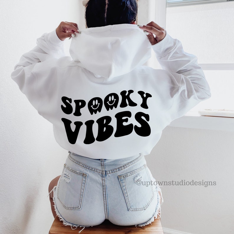 May include: White hoodie with black text that says "Spooky Vibes" in a retro font. There are three ghost faces with smiling eyes above the text.