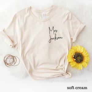 May include: A soft cream colored t-shirt with the text "Mrs. Jackson" in black script on the front.