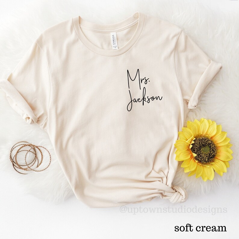 May include: A soft cream colored t-shirt with the text "Mrs. Jackson" in black script on the front.
