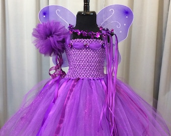 May include: Purple fairy tutu costume with glitter tulle skirt, purple satin ribbon, and butterfly wings. The costume also includes a purple flower and a wand.