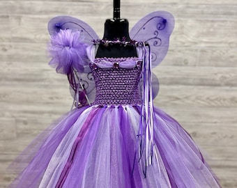 May include: A purple fairy costume featuring a crochet bodice with a tulle skirt in shades of light and dark purple. The outfit includes sheer purple wings with black swirl patterns, a star-adorned neckline, and a matching wand with a purple pom-pom.