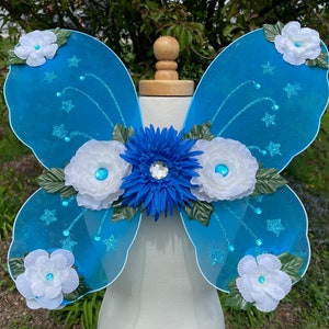 May include: Blue fairy wings with white flowers and a blue flower in the center. The wings are decorated with glitter and stars.