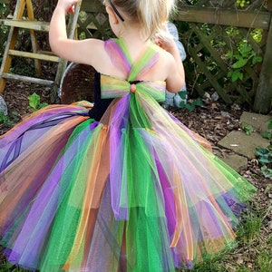 K&ouml;nnte beinhalten: Ein kleines Kind tr&auml;gt ein schwarzes Hexenkost&uuml;m mit einem regenbogenfarbenen T&uuml;llrock und einem schwarzen Hexenhut. Das Kind steht in einem grasbewachsenen Bereich mit einem Holzzaun im Hintergrund.
