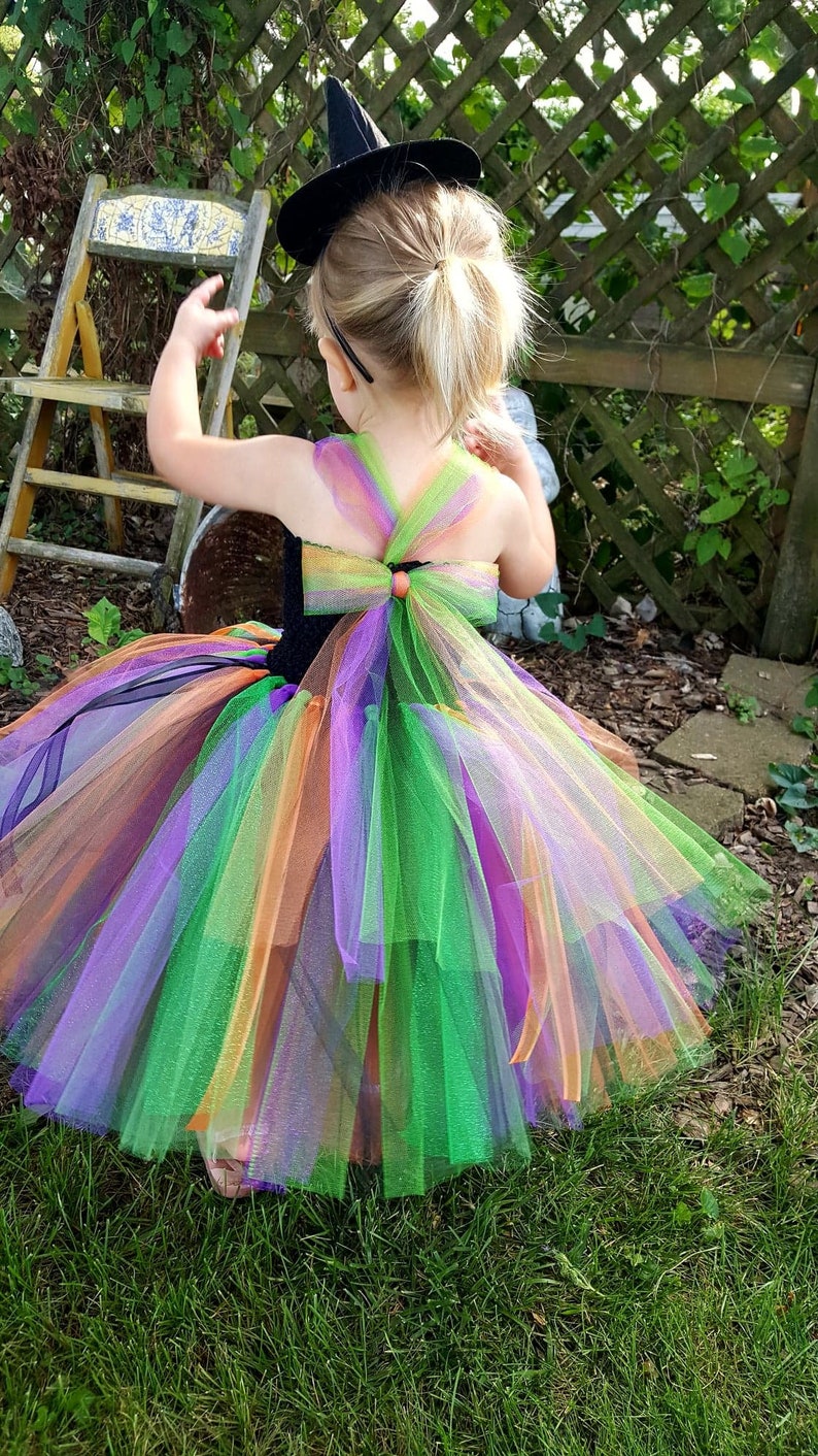 K&ouml;nnte beinhalten: Ein kleines Kind tr&auml;gt ein schwarzes Hexenkost&uuml;m mit einem regenbogenfarbenen T&uuml;llrock und einem schwarzen Hexenhut. Das Kind steht in einem grasbewachsenen Bereich mit einem Holzzaun im Hintergrund.