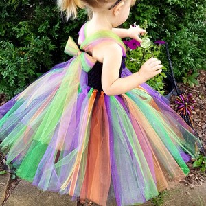 K&ouml;nnte beinhalten: Ein junges M&auml;dchen tr&auml;gt einen bunten T&uuml;ll-Tutu mit einem schwarzen Oberteil und einem schwarzen Hexenhut. Der Tutu hat gr&uuml;ne, lila, orange und gelbe Schichten.