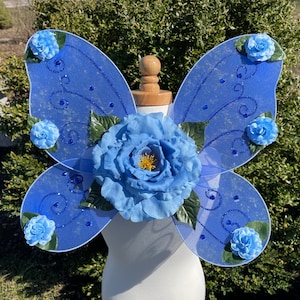 May include: Blue fairy wings with a large blue flower in the centre and smaller blue flowers on the edges. The wings are made of sheer fabric with glitter and blue swirls.