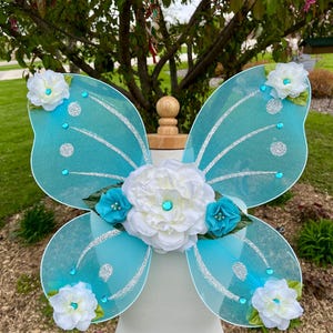 May include: Blue butterfly fairy wings with white and turquoise flowers. The wings are made of sheer fabric with glitter accents.