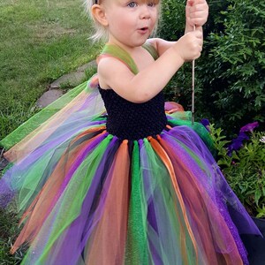 K&ouml;nnte beinhalten: Ein kleines Kind verkleidet als Hexe f&uuml;r Halloween. Das Kind tr&auml;gt einen schwarzen Hexenhut mit gr&uuml;nen und orangefarbenen Akzenten, einen schwarzen und mehrfarbigen T&uuml;ll-Tutu und ein schwarzes &auml;rmelloses Oberteil. Das Kind h&auml;lt einen Zauberstab mit einer durchsichtigen Glaskugel oben.