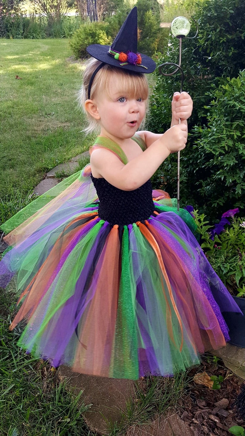 K&ouml;nnte beinhalten: Ein kleines Kind verkleidet als Hexe f&uuml;r Halloween. Das Kind tr&auml;gt einen schwarzen Hexenhut mit gr&uuml;nen und orangefarbenen Akzenten, einen schwarzen und mehrfarbigen T&uuml;ll-Tutu und ein schwarzes &auml;rmelloses Oberteil. Das Kind h&auml;lt einen Zauberstab mit einer durchsichtigen Glaskugel oben.