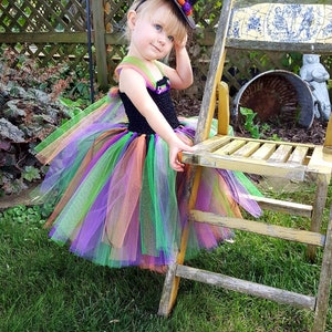 K&ouml;nnte beinhalten: Ein kleines Kind, das sich f&uuml;r Halloween als Hexe verkleidet hat. Sie tr&auml;gt einen schwarzen und gr&uuml;nen Tutu mit orangefarbenen und violetten Akzenten. Sie tr&auml;gt auch einen schwarzen Hexenhut mit einer gr&uuml;nen und orangefarbenen Blume oben drauf. Das Kind sitzt auf einem Holzstuhl in einem grasbewachsenen Bereich.