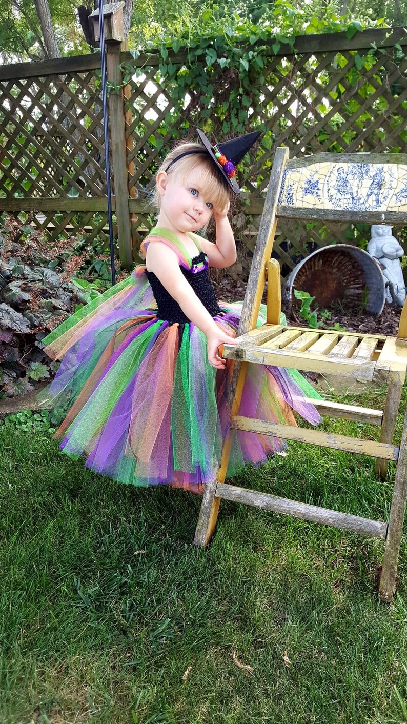 K&ouml;nnte beinhalten: Ein kleines Kind, das sich f&uuml;r Halloween als Hexe verkleidet hat. Sie tr&auml;gt einen schwarzen und gr&uuml;nen Tutu mit orangefarbenen und violetten Akzenten. Sie tr&auml;gt auch einen schwarzen Hexenhut mit einer gr&uuml;nen und orangefarbenen Blume oben drauf. Das Kind sitzt auf einem Holzstuhl in einem grasbewachsenen Bereich.