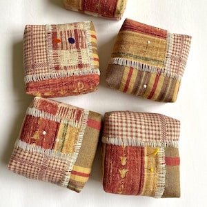 May include: Five small, square, patchwork pincushions in shades of brown, red, and cream. Each pincushion has a different pattern and is decorated with decorative stitching. Some pincushions have pins inserted into them.