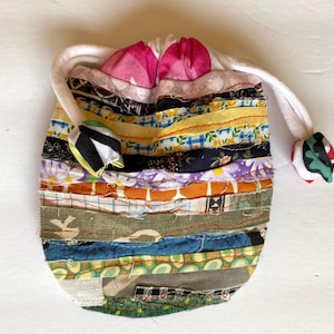 May include: A handmade drawstring pouch crafted from various fabric scraps in a patchwork design. The pouch features a white drawstring and is adorned with pink and patterned fabric accents. The overall color scheme includes a mix of blues, yellows, and greens.