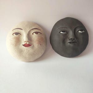 May include: Two circular wall plaques featuring sculpted human faces. One plaque is off-white with painted brown eyebrows, dark eyes, and red lips. The other plaque is dark grey with sculpted facial features, including eyes and a mouth.