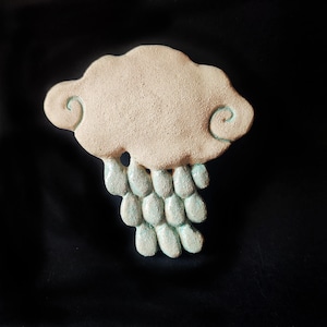 Puede incluir: Un adorno de pared de cerámica texturizada con forma de nube con extremos en espiral y gotas de lluvia colgantes. La nube es de color blanco roto mate, mientras que los elementos de lluvia en forma de lágrima tienen un esmalte turquesa brillante.