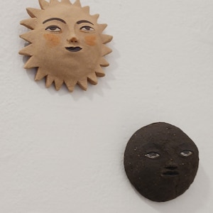 Puede incluir: Dos placas de cerámica para pared con rostros. La pieza superior es un sol de color tostado claro con rayos puntiagudos, ojos pintados y mejillas naranjas. La pieza inferior es una luna de color marrón oscuro, texturizada, redondeada y con ojos pintados.