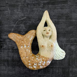 May include: A ceramic mermaid figurine with a brown and white tail and a white torso. The mermaid has her arms raised above her head and her eyes are closed.