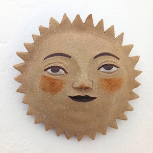 Op de afbeelding: Een ronde keramische zon-muurplaat met een getextureerde beige afwerking. De zon heeft een handgeschilderd gezicht met donkerbruine wenkbrauwen, ogen en lippen, samen met twee oranje-achtige cirkelvormige vlekken op de wangen.