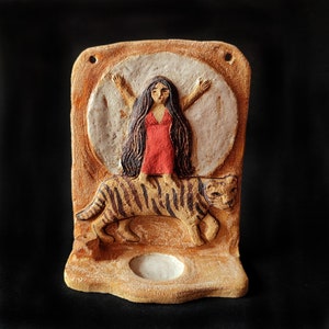 Puede incluir: Una placa de pared rectangular de arcilla con un relieve de una persona con cabello largo y oscuro que viste un vestido rojo, de pie sobre un tigre. La figura está colocada contra un gran disco circular blanco. Una pequeña hendidura está en la base.